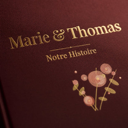 Macro de la couverture bordeaux « Marie & Thomas · Notre Histoire » : dorure et bouquet aquarelle sur lin